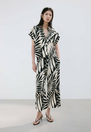 Žena má na sebe čierno-bielu košeľu s krátkym rukávom so vzorom listov a široké nohavice s čiernymi pásikmi na podpätkoch, stojí pri jednoduchej stene.