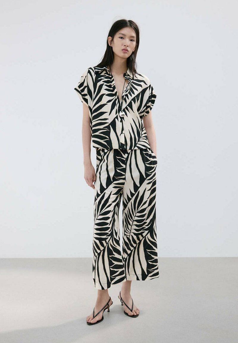 Femme portant une chemise à manches courtes assortie avec un motif de feuilles noir et blanc et un pantalon à jambes larges, avec des talons noirs à brides, debout contre un mur uni.