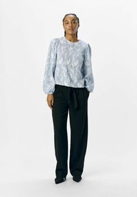 Blusa bordada de color azul claro con patrones florales, de mangas largas y puños fruncidos, combinada con pantalones negros de pierna ancha y zapatos negros de punta.