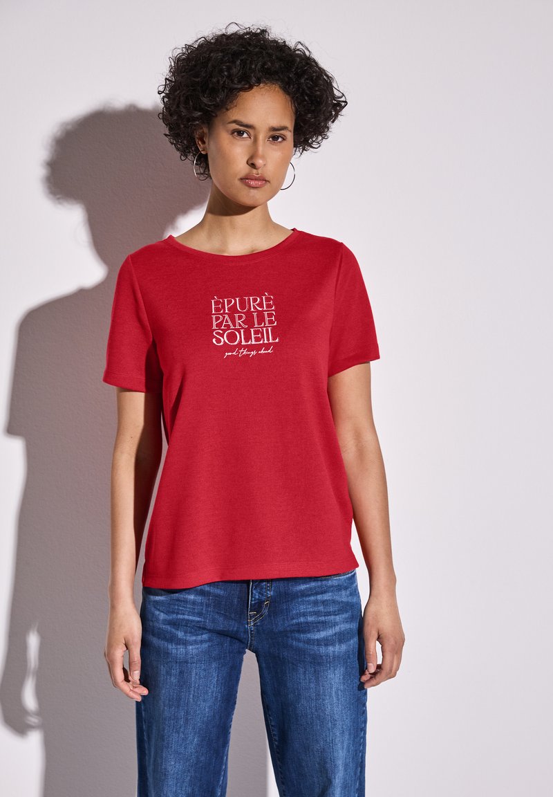 T-shirt rouge à manches courtes avec un col rond et un graphique en texte blanc "ÉPURÉ PAR LE SOLEIL" sur le devant, associé à un jean en denim bleu.