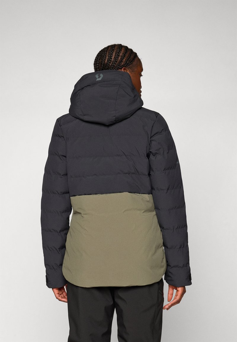 Person mit geflochtenem Haar, die eine schwarze und olivgrüne Kapuzenjacke trägt und mit dem Rücken zu einem schlichten hellen Hintergrund steht.