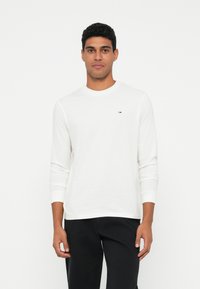 Homem de pé contra um fundo branco, vestindo uma camisa de manga longa branca com um pequeno logotipo no peito e calças pretas, virado para a frente.