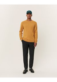 Mosterdgele gebreide trui met een ronde hals, gecombineerd met donkergrijze broek en zwarte schoenen. Het model draagt een donkergroene pet.