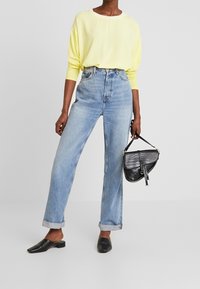 Person i en lys gul langærmet bluse, højtaljede lyseblå jeans med opslag, sorte slip-on sko, holder en sort tekstureret håndtaske.