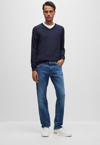 Jūrų karo ilgos rankovės V-formos megztinis užsidėtas ant balto marškinėlio, derinamas su šviesiai mėlynomis džinsinėmis kelnėmis ir baltomis sportinėmis bateliais su juodais ir raudonais akcentais.