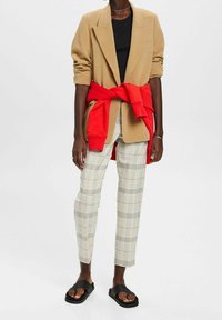 Beige blazer met verlaagde schouders, zwart t-shirt, rode sweatshirt om de taille geknoopt, geruite broek met zwarte en rode lijnen, en zwarte sandalen.