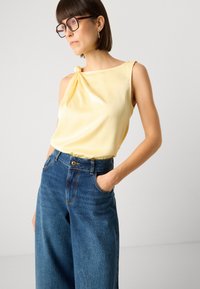 Top sem mangas de cetim amarelo com detalhe em nó no ombro, combinado com jeans de ganga azul de perna larga e cintura alta.