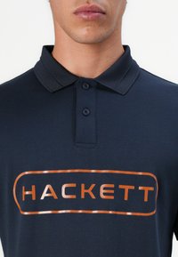 Sötétkék pólóing, puha anyagból készült, elöl réz színű "HACKETT" logóval. Klasszikus gallérral és két gombbal kiegészítve.