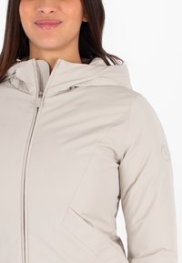 Giacca beige con cappuccio, tessuto liscio, chiusura con zip davanti, cuciture diagonali sottili ai lati, logo circolare sulla manica sinistra.