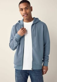 Sudadera con cremallera de color azul claro, con tejido texturizado, cordones y puños acanalados. Llevada sobre una camiseta blanca, combinada con jeans azules.
