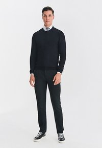 Czarny sweter z dzianiny, biała koszula z kołnierzykiem i czarne eleganckie spodnie. Szare sneakersy z białymi podeszwami. Gładkie tło, neutralne oświetlenie.