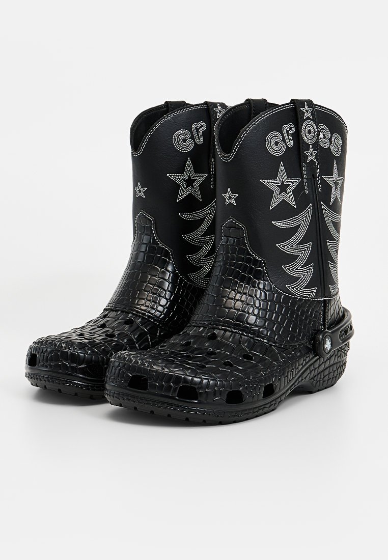 Botas de vaquero de plástico negro con textura de cocodrilo, marca grabada, patrones de estrellas y árboles, y detalles laterales perforados.