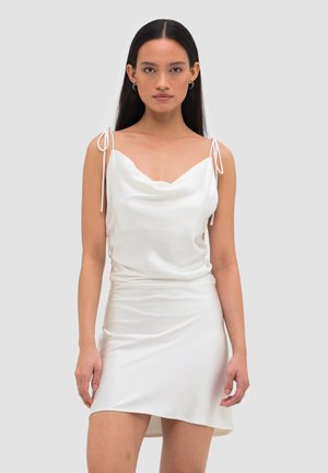 Kvinde med langt mørkt hår iført en hvid satin minikjole med bindbare skulderstropper, stående mod en ensfarvet lys baggrund.