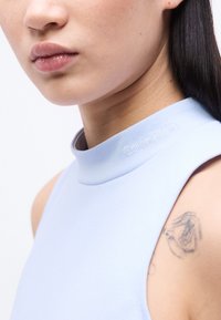 Κοντινό πλάνο ενός ατόμου που φοράει αμάνικη γαλάζια μπλούζα με τη λέξη «Calvin Klein» κεντημένη στον γιακά και ένα τατουάζ με μαύρο μελάνι στον πάνω βραχίονα.