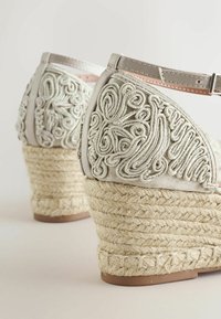 Espadrilles compensées beige avec talons en corde tressée et broderie en relief complexe sur la tige, dotées de brides à la cheville avec boucles.