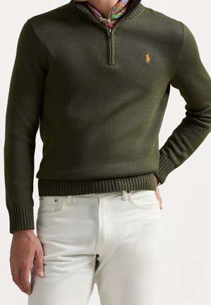 Homme portant un pull en tricot kaki à demi-fermeture éclair avec un petit logo sur la poitrine, une écharpe multicolore, et un pantalon blanc cassé, main dans la poche.