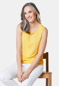 Top giallo senza maniche realizzato in tessuto morbido, abbinato a pantaloni bianchi. La modella è seduta su una sedia di legno con uno sfondo neutro.
