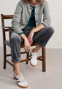 Šedý pletený cardigan s kulatým výstřihem, dřevěnými knoflíky, zelenou košilí pod ním, šedými kordurovými kalhotami a bílými teniskami se zelenými pruhy.