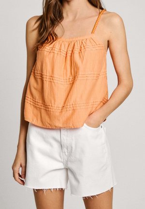 Woman wearing an orange sleeveless top with braided straps and white high-waisted denim shorts, hand resting in pocket.