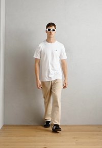Hvid t-shirt med korte ærmer, beige bukser og sorte sko. Modellen har hvide solbriller på. Baggrunden viser en lys grå væg og et trægulv.
