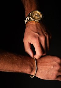 Orologio d'oro con una lunetta incoronata di gioielli a un polso, e un sottile bracciale d'oro che viene aggiustato sull'altro polso, entrambi su uno sfondo scuro.