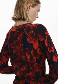 Piros és fekete virágmintás pulóver hosszú ujjal, bordás nyakkivágással és gombos részletekkel a hátán. Puha szövet textúra.