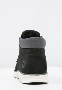 Svart mocka ankelsko med en grå vadderad krage, vit sula och präglad logotyp på utsidan. Slät textur och elegant design.