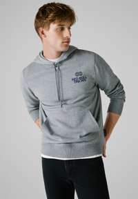 Felpa grigia con tasca frontale, cappuccio con cordino e logo navy "Red Bull Racing". Presenta polsini e orlo a coste, indossata sopra una maglietta bianca.