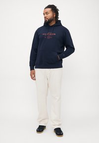 Felpa blu navy con tasca frontale, caratterizzata da un logo rosso. Abbinata a pantaloni chiari e sneakers nere con suole in gomma.