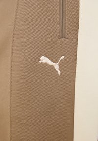 Barna sportnadrág, texturált felülettel, fehér Puma logóval és kontrasztos krémszínű oldalsó panellel. Zárható részlet látható.