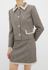 Brązowy i beżowy zestaw w desenie houndstooth, składający się z marynarki z kołnierzykiem zapinanej na guziki oraz dopasowanej spódnicy, z kontrastowymi wykończeniami i subtelnymi ozdobami.