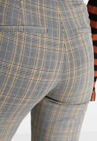 Grå och svart rutiga byxor med orange detaljer, med en mjuk textur och bakfickor. Mönstret är en blandning av rutor och linjer.