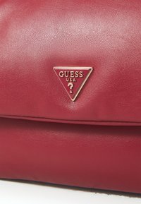 Borsa in pelle rossa con texture, con il logo triangolare dorato di Guess USA che presenta un punto interrogativo al centro.