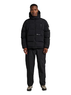 Mand iført sort dunjakke med hætte, sorte bukser og grå vandresko, stående mod en hvid baggrund.