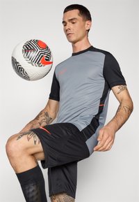 Sportskjorte og -shorts i grått og svart, med røde detaljer og en logo, med en teksturert fotball som balanseres.
