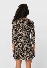 Mujer con cabello rizado y largo, vistiendo un vestido de leopardo de manga larga y corto, de pie de espaldas frente a un fondo liso.