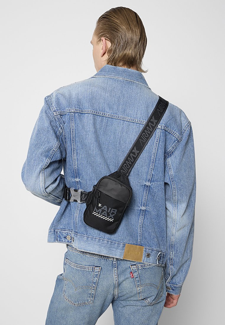 Persona vestida con una chaqueta de mezclilla azul clara y jeans, llevando una pequeña bolsa cruzada negra de Air Max con una correa ancha de marca sobre la espalda.