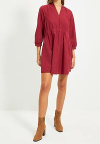 Rotes Baumwollkleid mit Dreiviertelärmeln, elastischem Bund und V-Ausschnitt. Tragen mit beigen Wildleder-Ankle-Boots mit Blockabsatz.