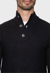 Sweter zapinany na guziki w ciemnym granacie, z fakturowanym wzorem rybiej łuski, wyposażony w wysoki kołnierz i dwa wyraźne guziki przy szyi.