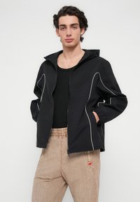 Μαύρο μπουφάν με λευκές λεπτομέρειες περιγράμματος, κλείσιμο με φερμουάρ και κουκούλα. Φορεμένο πάνω από μαύρο πουκάμισο, συνδυασμένο με ανοιχτόχρωμο παντελόνι με ελαστική μέση.