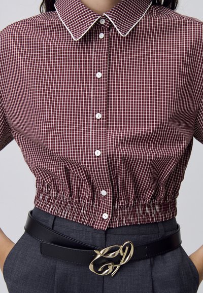 Camisa a cuadros marrón con cuello blanco, cintura elástica ceñida y botones en la parte frontal, combinada con un cinturón negro con un acento dorado.