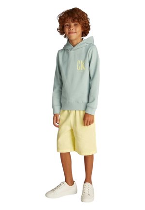 Sudadera con capucha de color azul claro con un logo amarillo, combinada con pantalones cortos amarillos y zapatillas blancas. Material de algodón, diseño casual, corte relajado.