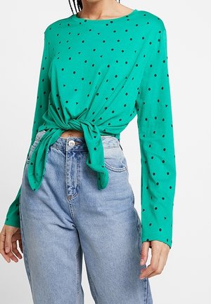 Grøn bluse med lange ærmer og sorte prikker, med et frontknyttet design. Kombineret med højtaljede blå denimjeans.
