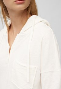 Sudadera blanca de textura con un bolsillo frontal, cordones y cuello abotonado. Presenta un corte relajado y un material suave.