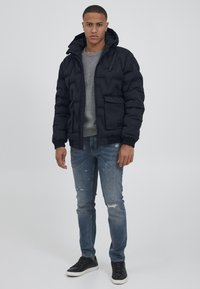Pufferjakke i mørk marineblå, med glidelås foran, hette og ribbestrikkede mansjetter. Brukt over en grå genser og kombinert med slitte blå jeans og mørke sneakers.