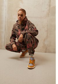 Sada hnědé mikiny a tepláků s kamuflážním vzorem a červenými a zelenými akcenty. Šedé a oranžové vysoké tenisky s černými detaily.