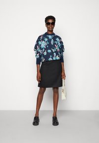 Suéter de patrón floral en azul marino, falda negra, zapatos negros de slip-on y bolso blanco, llevados por una modelo con cabello corto y gafas de sol.