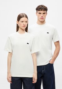 Λευκά βαμβακερά μπλουζάκια με άνετη εφαρμογή. Κάθε ένα φέρει ένα μικρό μαύρο λογότυπο στο αριστερό μέρος του στήθους. Φοριούνται με σκούρο τζιν παντελόνι.