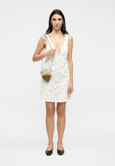 Γυναίκα που φοράει άσπρο δαντελένιο αμάνικο φόρεμα, μαύρες μπαλαρίνες και κρατά μια μικρή τσάντα με χάντρες σε σχήμα βελανιδιού στον ώμο της.