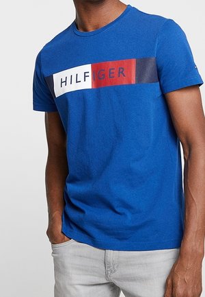 Homme portant un T-shirt bleu avec le texte "HILFIGER" en blocs blanc, rouge et marine, associé à un pantalon gris clair, main dans la poche.